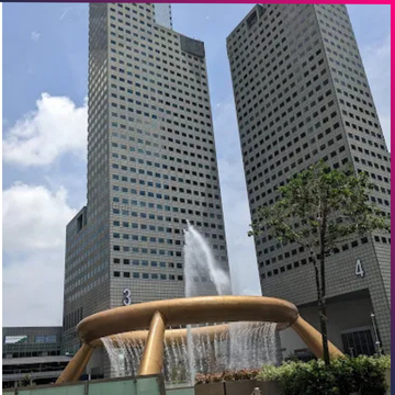 Two tall office buildings stand behind a circular fountain with water spraying upwards, surrounded by trees and a clear sky, setting the stage for Memcyco at Global Anti-Scam Summit Asia 2025.